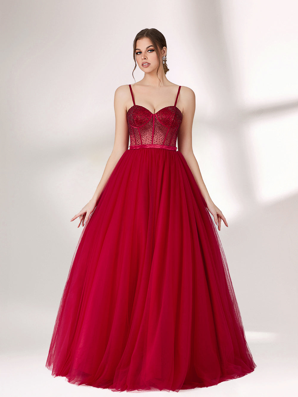 A woman stands against a light background, wearing a deep red evening gown. It features thin spaghetti straps, a sweetheart neckline with a sequined bodice that has sheer, dotted detailing. The skirt is a voluminous, flowing tulle, creating an elegant and romantic look, perfect for formal events.5
