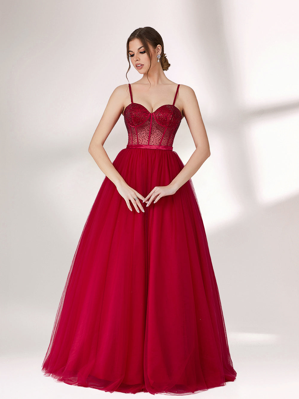 A woman stands against a light background, wearing a deep red evening gown. It features thin spaghetti straps, a sweetheart neckline with a sequined bodice that has sheer, dotted detailing. The skirt is a voluminous, flowing tulle, creating an elegant and romantic look, perfect for formal events.1