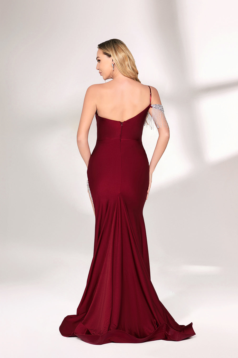 A woman with long, wavy blonde hair and a poised stance wears a deep - red satin mermaid gown. It has an asymmetrical neckline with one shoulder adorned by sparkling, dangling embellishments and a high side slit. Paired with simple heels, the outfit radiates elegance, perfect for formal events like galas.002