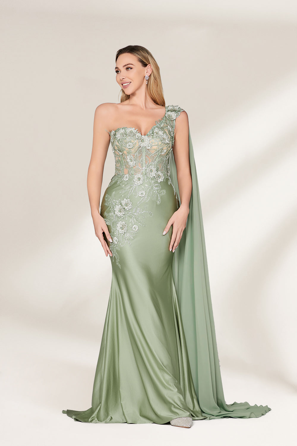 A woman stands against a light background, wearing a sage - green satin gown. It features a one - shoulder design with a flowing cape, floral lace embroidery on the bodice and skirt, and a mermaid silhouette. The elegant details and unique cape create a sophisticated look, perfect for formal events like galas or weddings.1