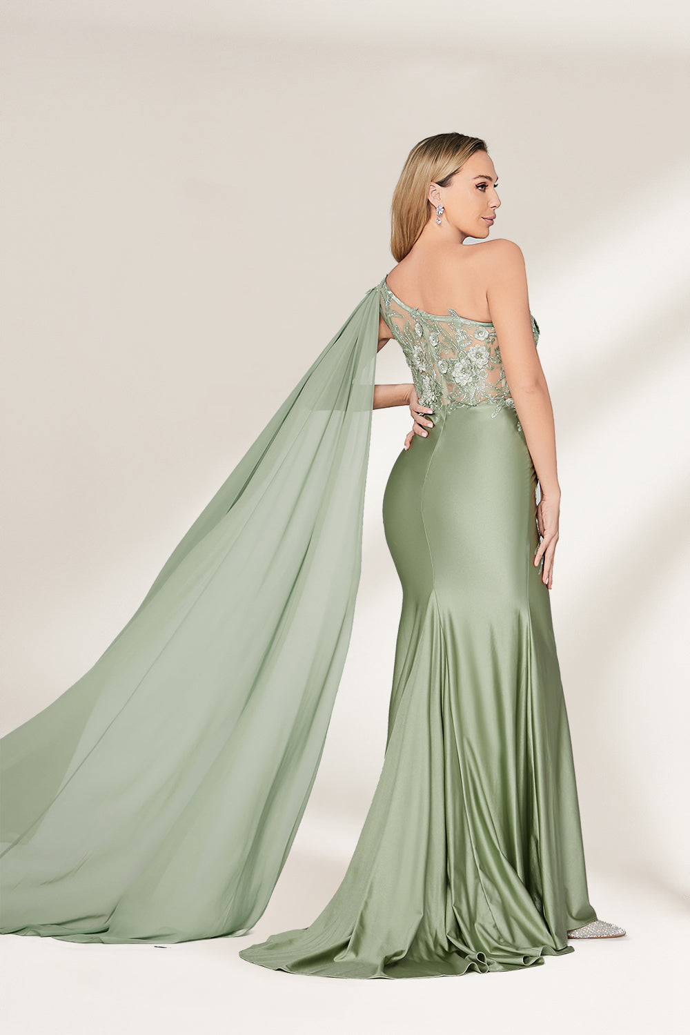 A woman stands against a light background, wearing a sage - green satin gown. It features a one - shoulder design with a flowing cape, floral lace embroidery on the bodice and skirt, and a mermaid silhouette. The elegant details and unique cape create a sophisticated look, perfect for formal events like galas or weddings.2