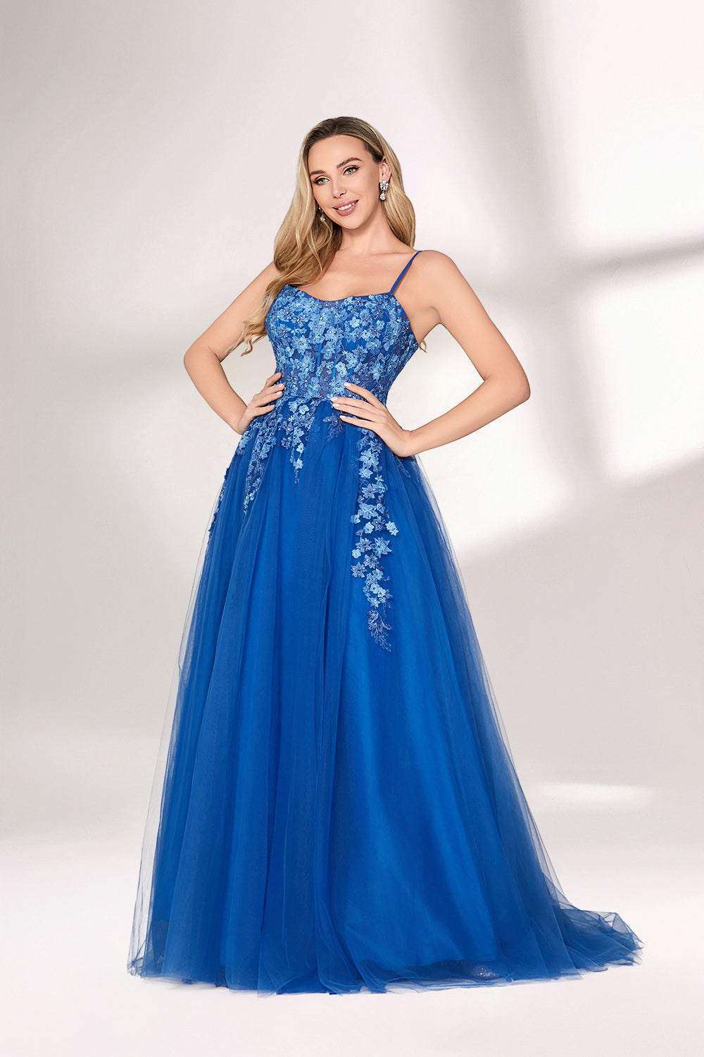 A woman stands against a light background, wearing a bright blue evening gown. It features thin spaghetti straps, a sweetheart neckline, and a bodice adorned with delicate floral lace embroidery. The voluminous tulle skirt flows elegantly, creating a romantic and glamorous look, perfect for formal events.3