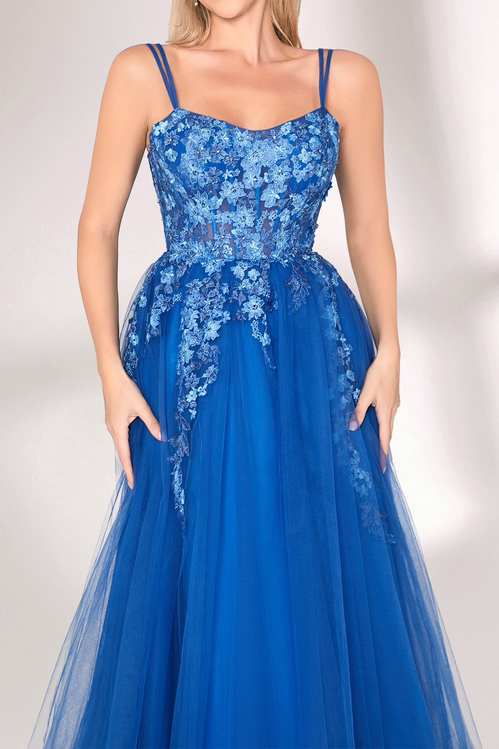 A woman stands against a light background, wearing a bright blue evening gown. It features thin spaghetti straps, a sweetheart neckline, and a bodice adorned with delicate floral lace embroidery. The voluminous tulle skirt flows elegantly, creating a romantic and glamorous look, perfect for formal events.5