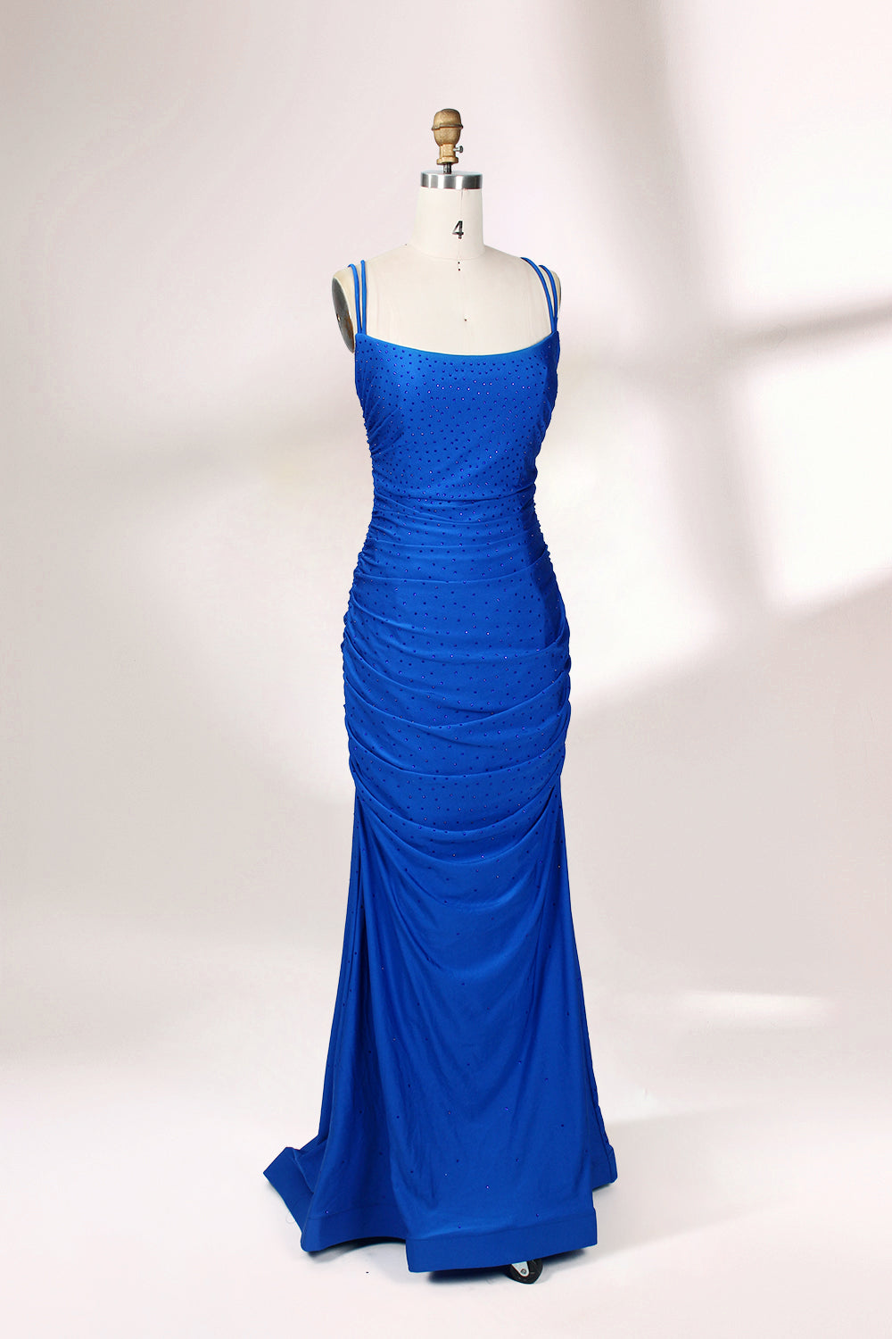 Photograph of a blue, floor-length evening gown displayed on a white dress form. The gown is sleeveless with a fitted bodice and a flared skirt, featuring thin shoulder straps and a textured, ruched design. The dress form is placed against a plain, off-white background, casting soft shadows.03