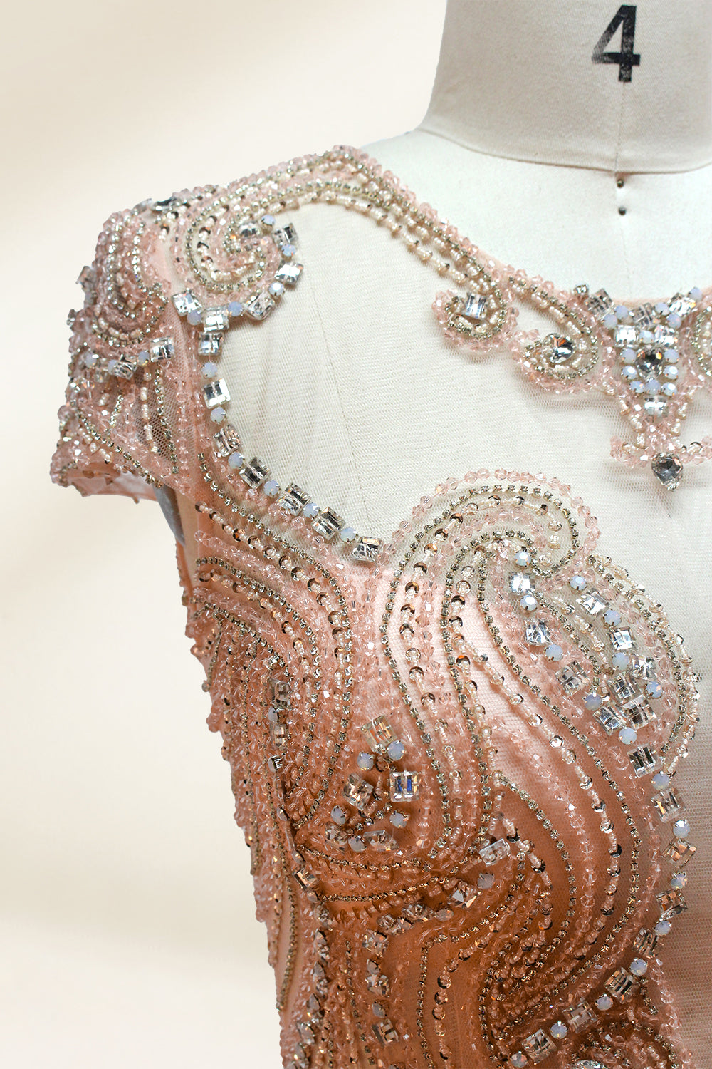 Photograph of an elegant, floor-length, peach-colored evening gown on a mannequin. The gown features intricate, sparkling beadwork on the bodice and cap sleeves, and a flowing, sheer skirt. The mannequin has a light beige background, with soft shadows highlighting the gown's details. The dress exudes luxury and sophistication.3132.4