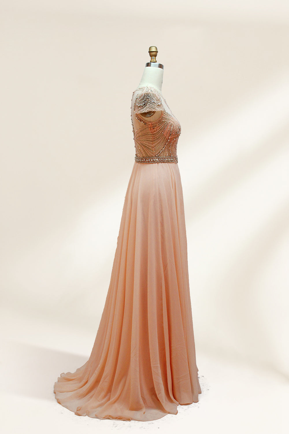 Photograph of an elegant, floor-length, peach-colored evening gown on a mannequin. The gown features intricate, sparkling beadwork on the bodice and cap sleeves, and a flowing, sheer skirt. The mannequin has a light beige background, with soft shadows highlighting the gown's details. The dress exudes luxury and sophistication.3132.5
