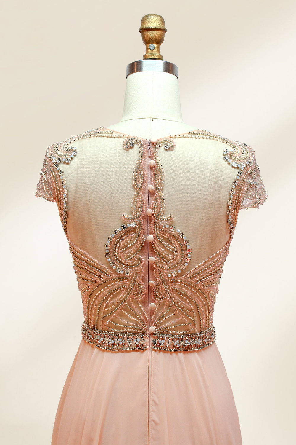 Photograph of an elegant, floor-length, peach-colored evening gown on a mannequin. The gown features intricate, sparkling beadwork on the bodice and cap sleeves, and a flowing, sheer skirt. The mannequin has a light beige background, with soft shadows highlighting the gown's details. The dress exudes luxury and sophistication.3132.6