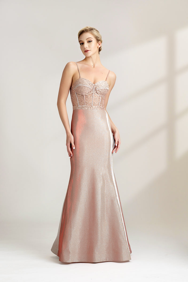 A photograph of a slender, fair-skinned woman with short blonde hair, wearing a floor-length, shimmering silver gown with a lace bodice and spaghetti straps. She stands confidently against a plain white background with soft, diffused light casting subtle shadows. The gown features a flattering, form-fitting silhouette.32547-01