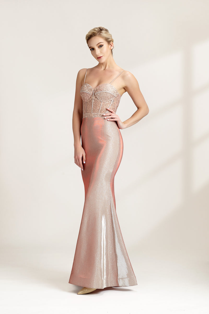 A photograph of a slender, fair-skinned woman with short blonde hair, wearing a floor-length, shimmering silver gown with a lace bodice and spaghetti straps. She stands confidently against a plain white background with soft, diffused light casting subtle shadows. The gown features a flattering, form-fitting silhouette.32547.03