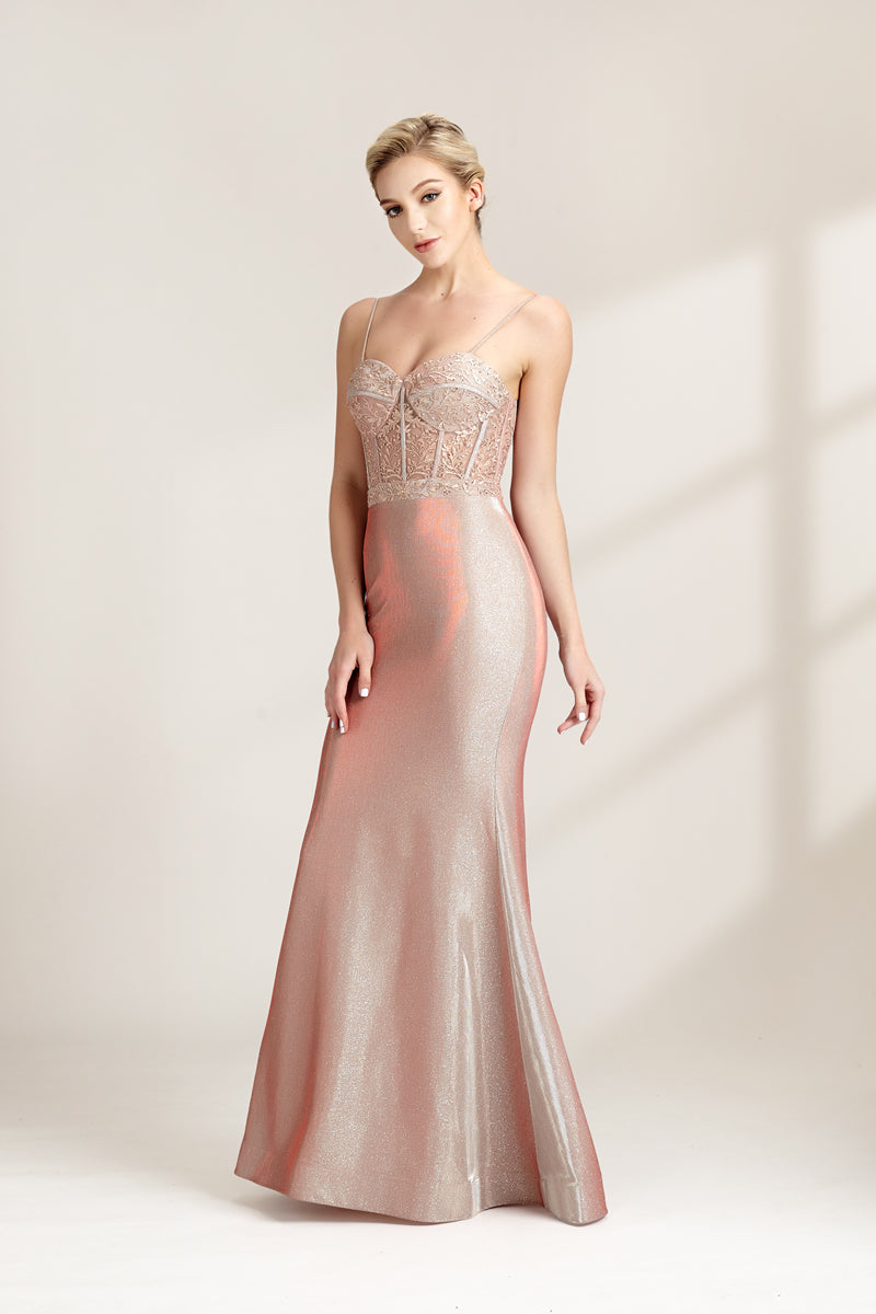 A photograph of a slender, fair-skinned woman with short blonde hair, wearing a floor-length, shimmering silver gown with a lace bodice and spaghetti straps. She stands confidently against a plain white background with soft, diffused light casting subtle shadows. The gown features a flattering, form-fitting silhouette.