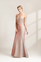 A photograph of a slender, fair-skinned woman with short blonde hair, wearing a floor-length, shimmering silver gown with a lace bodice and spaghetti straps. She stands confidently against a plain white background with soft, diffused light casting subtle shadows. The gown features a flattering, form-fitting silhouette.