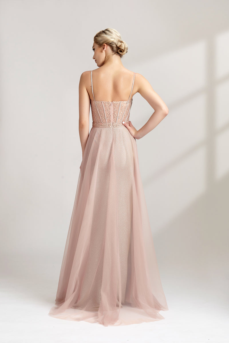 Photograph of a slim, fair-skinned woman with short blonde hair styled in a sleek updo. She wears a floor-length, light pink, sheer tulle gown with intricate lace detailing on the bodice. The gown has spaghetti straps and a sweetheart neckline. She stands against a plain white background, casting soft shadows. The overall aesthetic is elegant and sophisticated.32547b.02