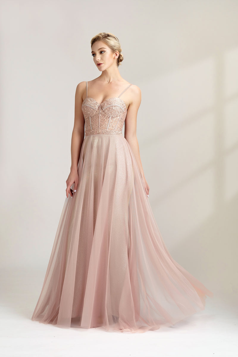 Photograph of a slim, fair-skinned woman with short blonde hair styled in a sleek updo. She wears a floor-length, light pink, sheer tulle gown with intricate lace detailing on the bodice. The gown has spaghetti straps and a sweetheart neckline. She stands against a plain white background, casting soft shadows. The overall aesthetic is elegant and sophisticated.32547b.04