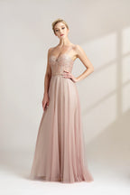 Photograph of a slim, fair-skinned woman with short blonde hair styled in a sleek updo. She wears a floor-length, light pink, sheer tulle gown with intricate lace detailing on the bodice. The gown has spaghetti straps and a sweetheart neckline. She stands against a plain white background, casting soft shadows. The overall aesthetic is elegant and sophisticated.32547b.05