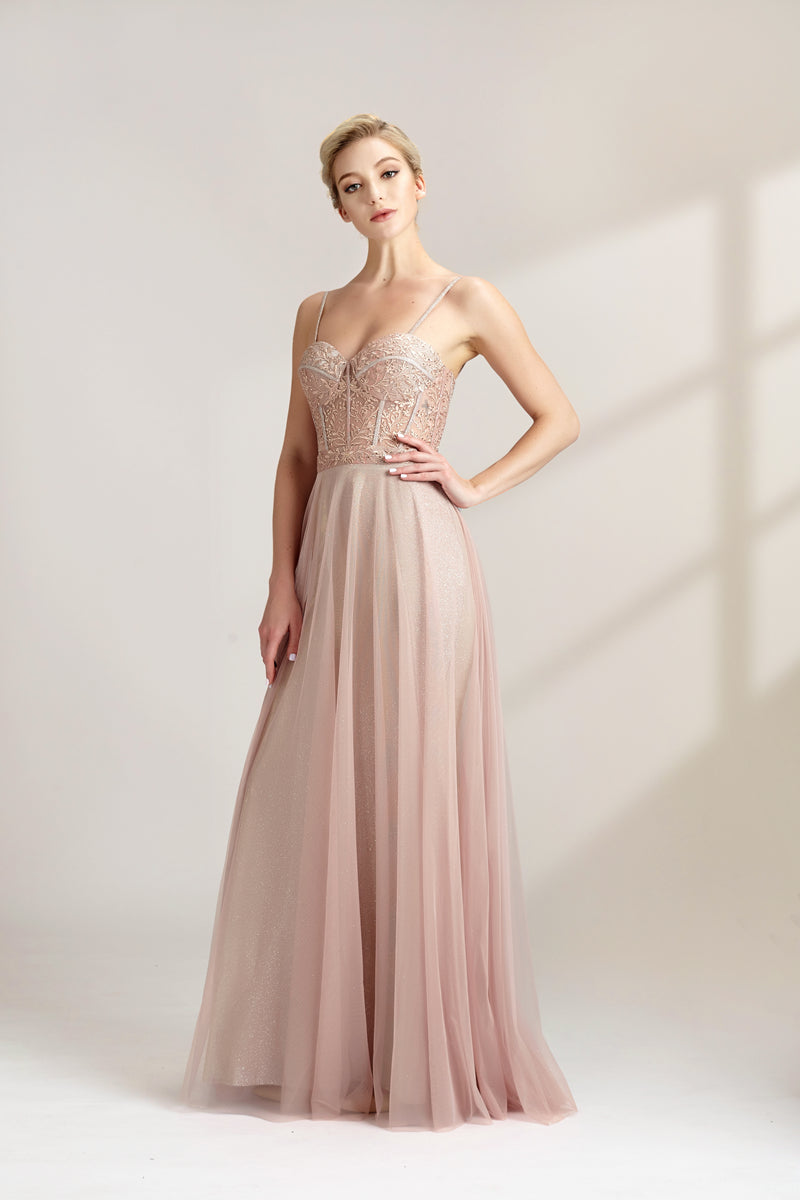 Photograph of a slim, fair-skinned woman with short blonde hair styled in a sleek updo. She wears a floor-length, light pink, sheer tulle gown with intricate lace detailing on the bodice. The gown has spaghetti straps and a sweetheart neckline. She stands against a plain white background, casting soft shadows. The overall aesthetic is elegant and sophisticated.32547b.05