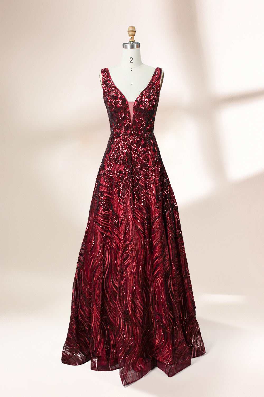 A photograph of a model dress on a dress form. The dress is a sleeveless, deep V-neck gown with a plunging neckline. It features a rich, deep red color with intricate, swirling black and red patterns, giving it a luxurious and elegant appearance. The background is a soft, neutral tone, with gentle shadows cast on the dress form.32625.1