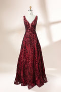 A photograph of a model dress on a dress form. The dress is a sleeveless, deep V-neck gown with a plunging neckline. It features a rich, deep red color with intricate, swirling black and red patterns, giving it a luxurious and elegant appearance. The background is a soft, neutral tone, with gentle shadows cast on the dress form.32625.1