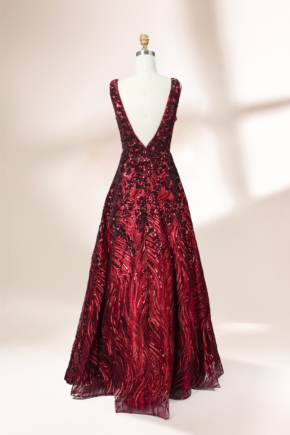 A photograph of a model dress on a dress form. The dress is a sleeveless, deep V-neck gown with a plunging neckline. It features a rich, deep red color with intricate, swirling black and red patterns, giving it a luxurious and elegant appearance. The background is a soft, neutral tone, with gentle shadows cast on the dress form.32625.2