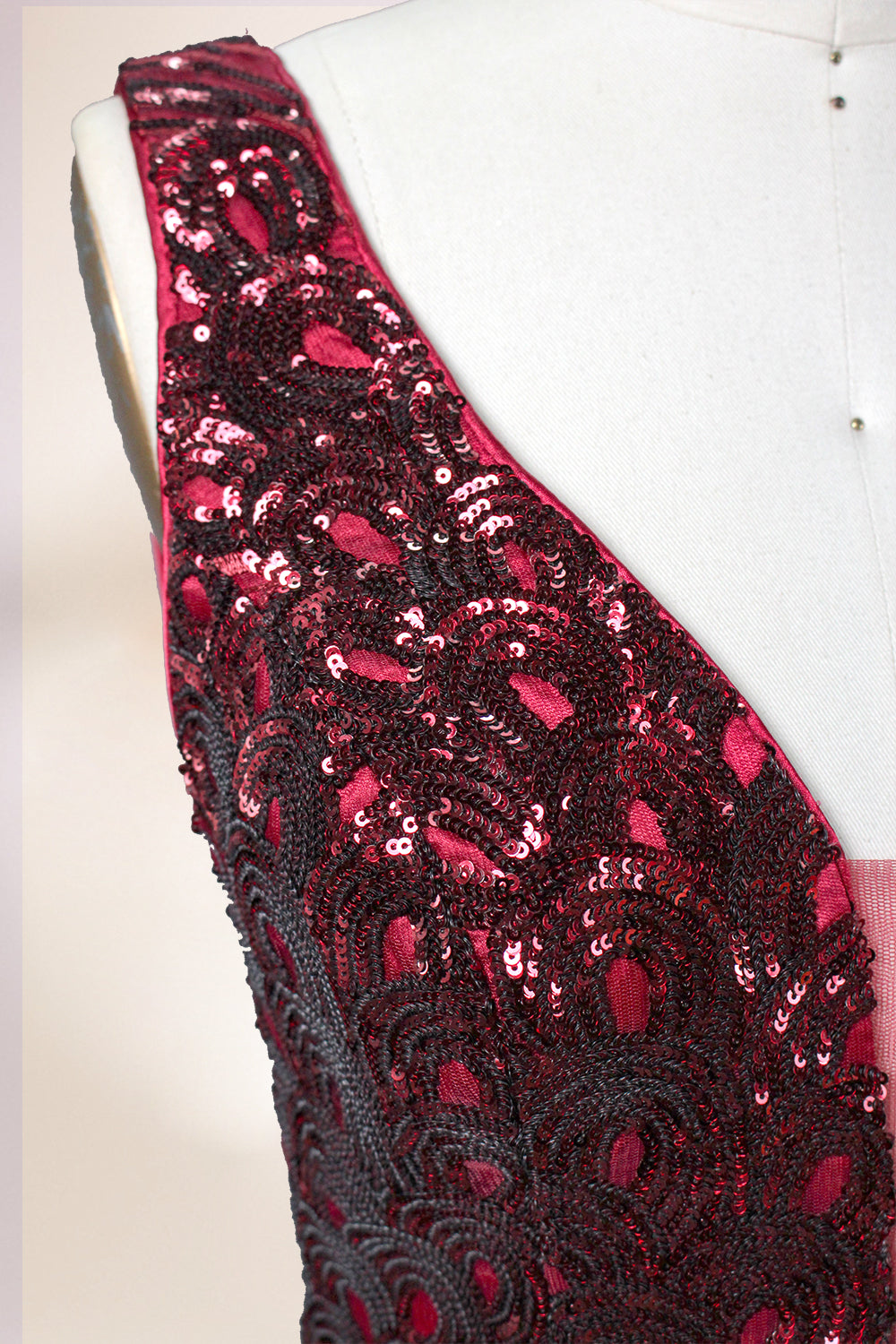 A photograph of a model dress on a dress form. The dress is a sleeveless, deep V-neck gown with a plunging neckline. It features a rich, deep red color with intricate, swirling black and red patterns, giving it a luxurious and elegant appearance. The background is a soft, neutral tone, with gentle shadows cast on the dress form.