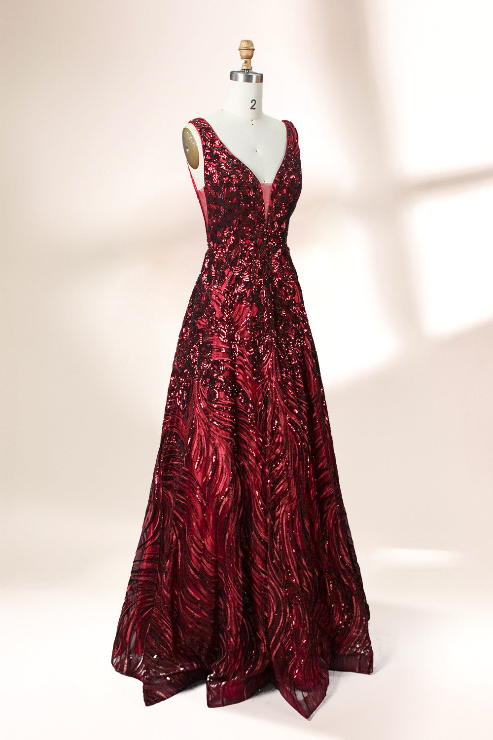 A photograph of a model dress on a dress form. The dress is a sleeveless, deep V-neck gown with a plunging neckline. It features a rich, deep red color with intricate, swirling black and red patterns, giving it a luxurious and elegant appearance. The background is a soft, neutral tone, with gentle shadows cast on the dress form.32625.6