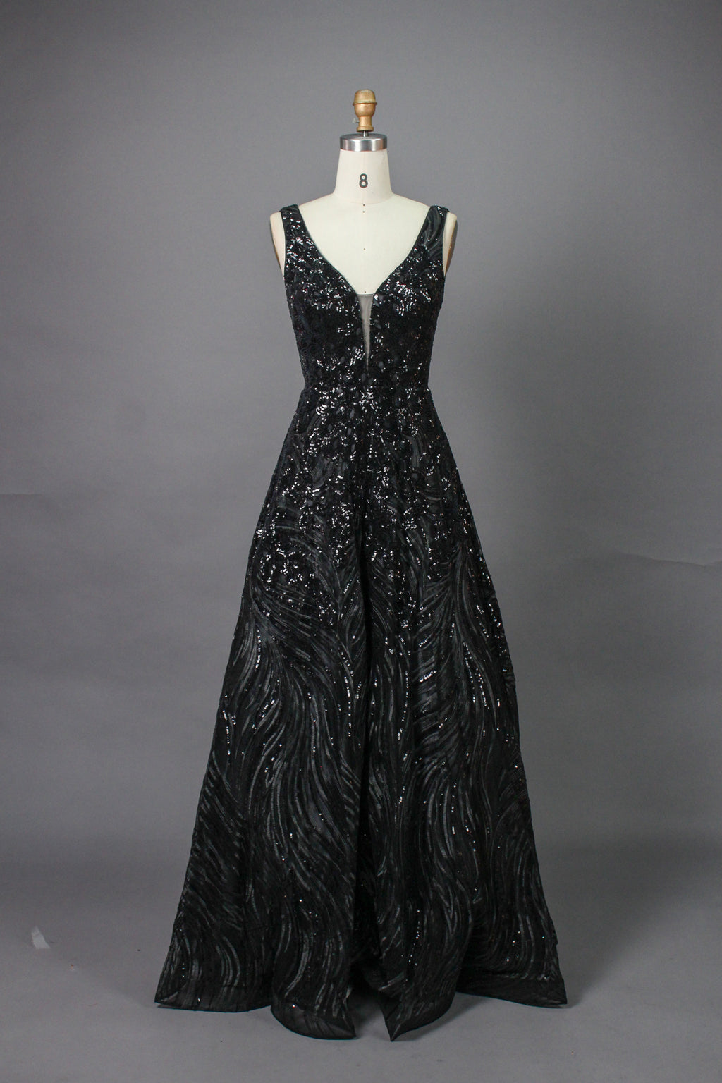 A photograph of a model dress on a dress form. The dress is a sleeveless, deep V-neck gown with a plunging neckline. It features a rich, deep red color with intricate, swirling black and red patterns, giving it a luxurious and elegant appearance. The background is a soft, neutral tone, with gentle shadows cast on the dress form.
