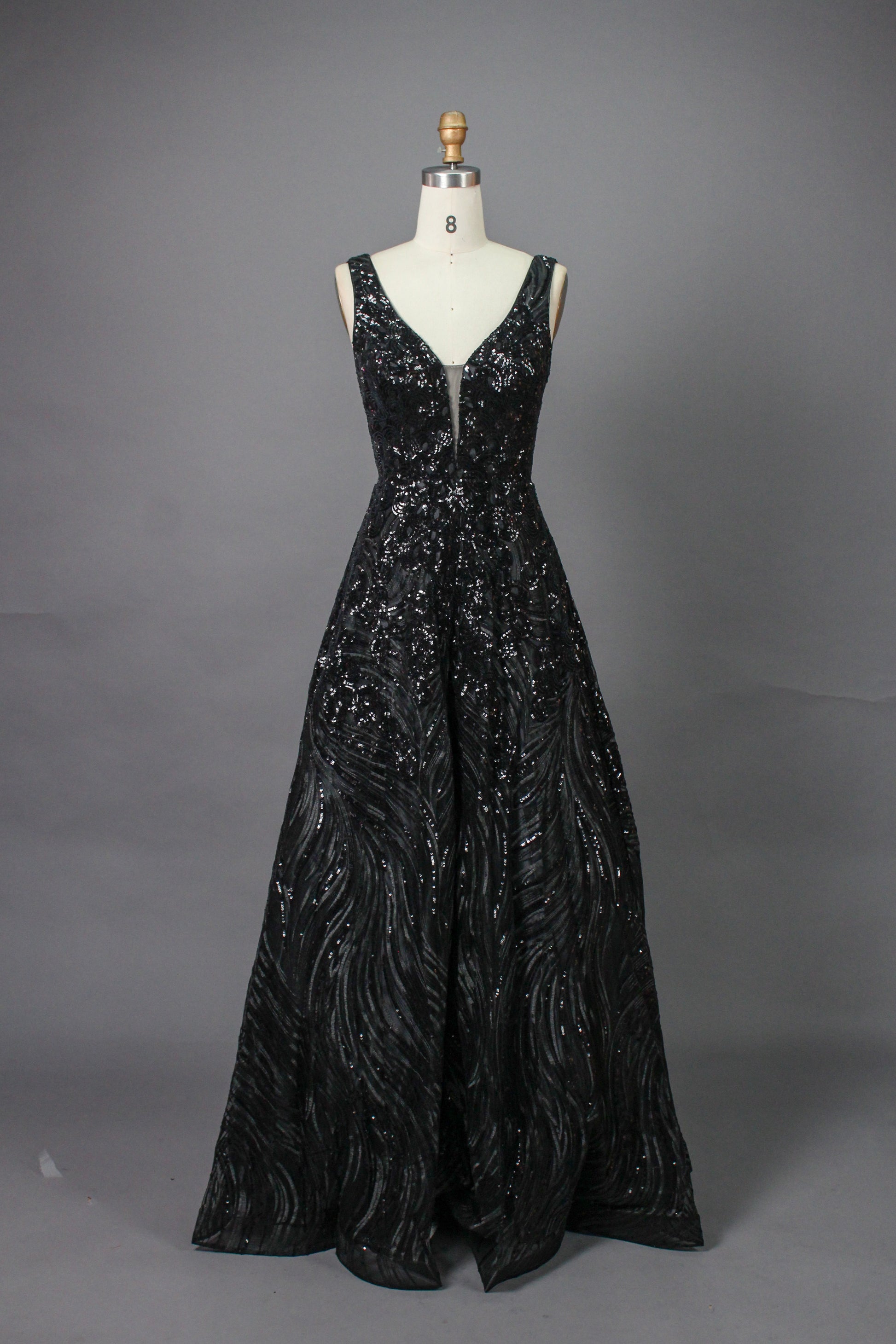 A photograph of a model dress on a dress form. The dress is a sleeveless, deep V-neck gown with a plunging neckline. It features a rich, deep red color with intricate, swirling black and red patterns, giving it a luxurious and elegant appearance. The background is a soft, neutral tone, with gentle shadows cast on the dress form.