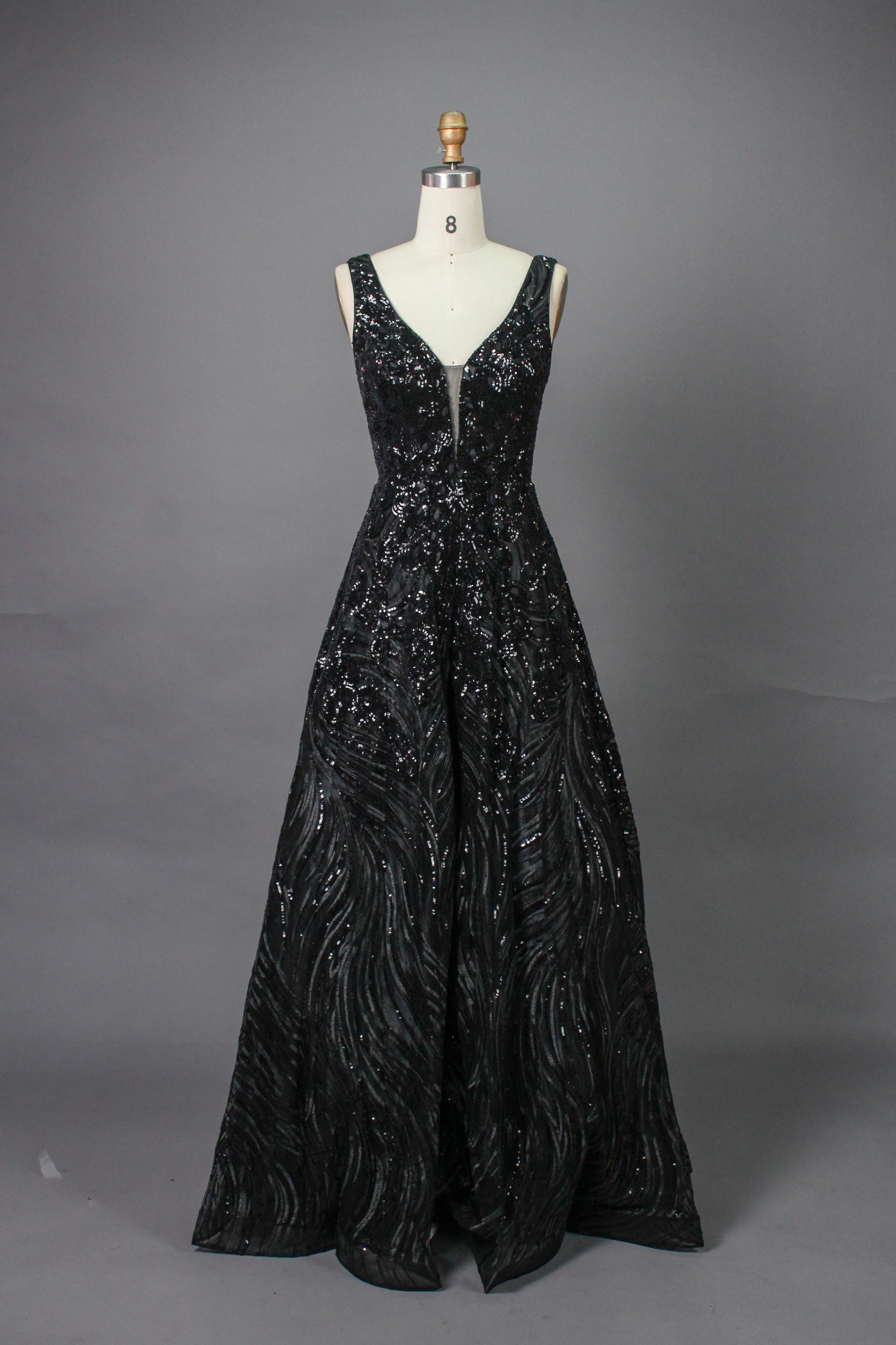 A photograph of a model dress on a dress form. The dress is a sleeveless, deep V-neck gown with a plunging neckline. It features a rich, deep red color with intricate, swirling black and red patterns, giving it a luxurious and elegant appearance. The background is a soft, neutral tone, with gentle shadows cast on the dress form.