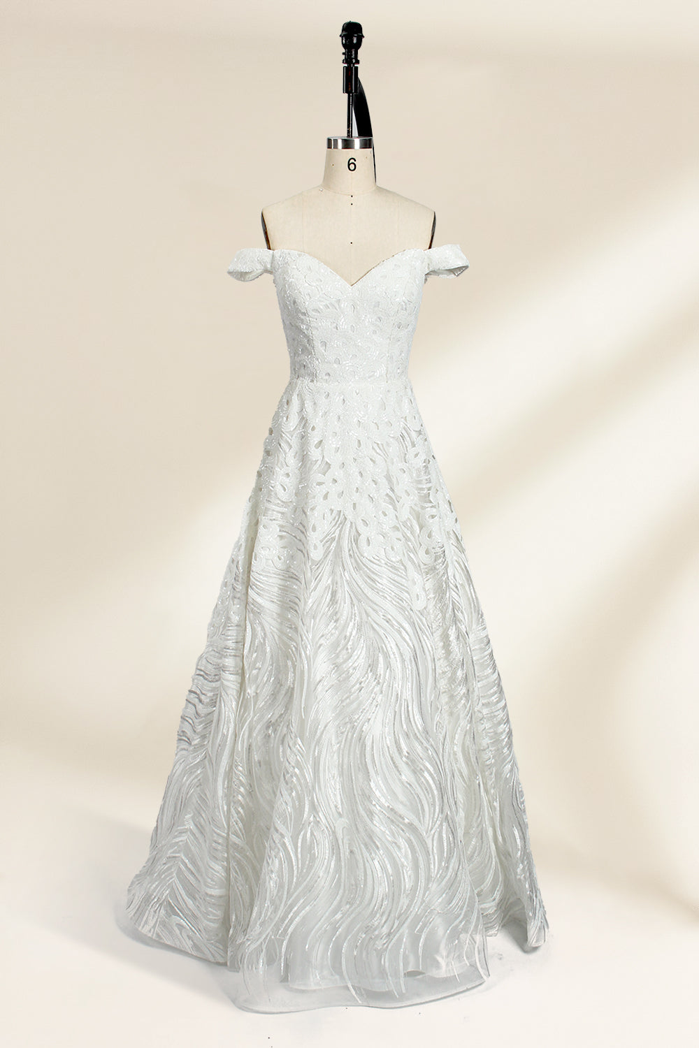 Photograph of a white, off-the-shoulder wedding gown displayed on a black dress form against a cream backdrop. The gown features intricate, swirling floral lace embroidery with a fitted bodice and a full, flared skirt, exuding elegance and sophistication. The dress form is centered, highlighting the gown's delicate detailing and flowing texture.32625b.1