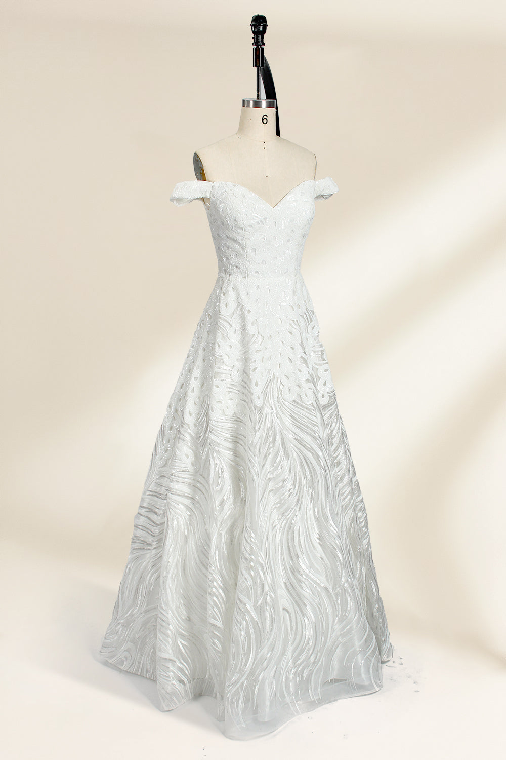 Photograph of a white, off-the-shoulder wedding gown displayed on a black dress form against a cream backdrop. The gown features intricate, swirling floral lace embroidery with a fitted bodice and a full, flared skirt, exuding elegance and sophistication. The dress form is centered, highlighting the gown's delicate detailing and flowing texture.32625b.3