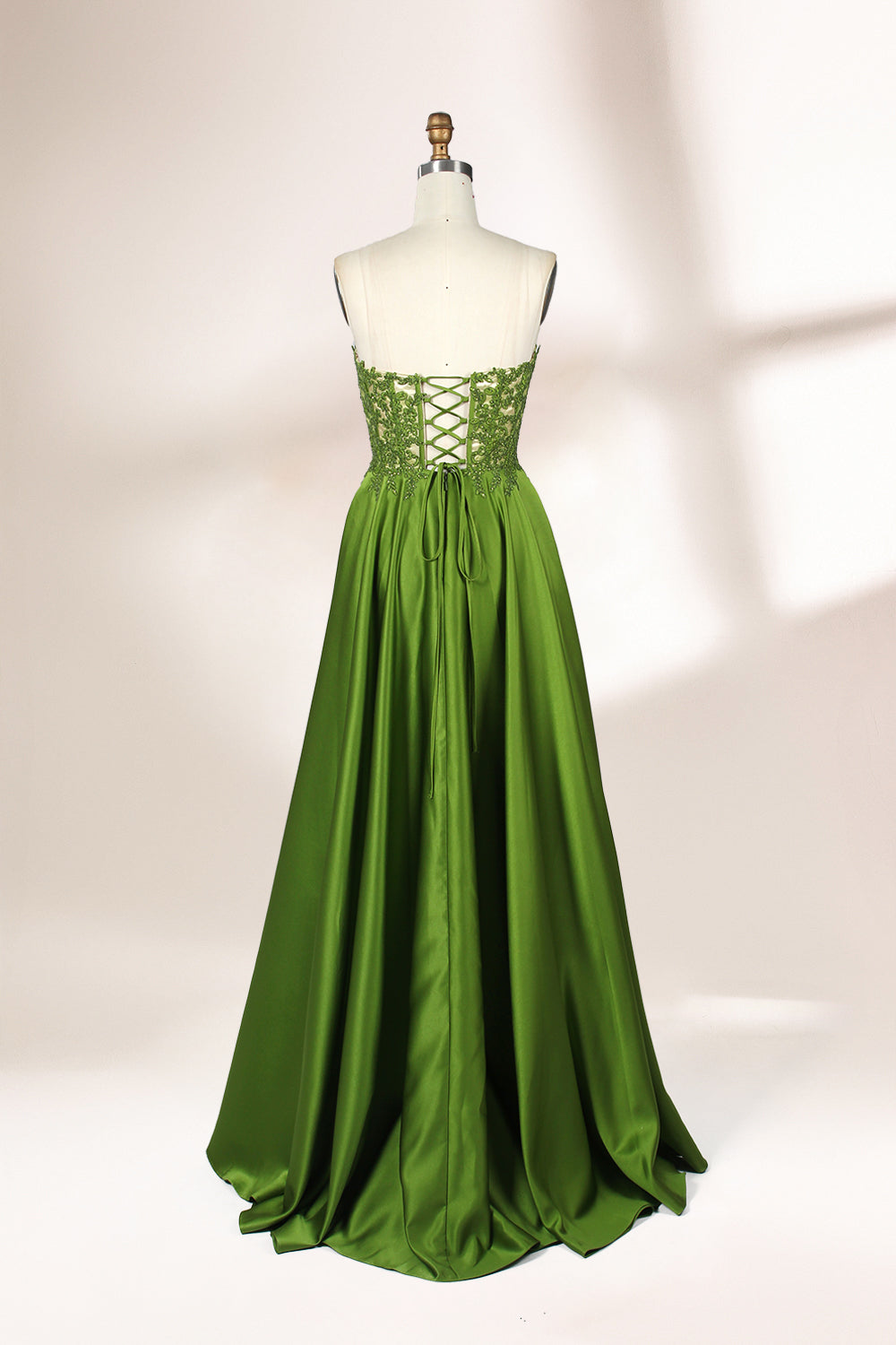 Photograph of an elegant, strapless gown displayed on a mannequin. The gown features a green, satin bodice adorned with intricate lace detailing and a full, floor-length skirt. The background is a soft, neutral gradient, enhancing the gown's vibrant green color. The mannequin is positioned centrally, with the gown's luxurious texture and refined craftsmanship prominently showcased.