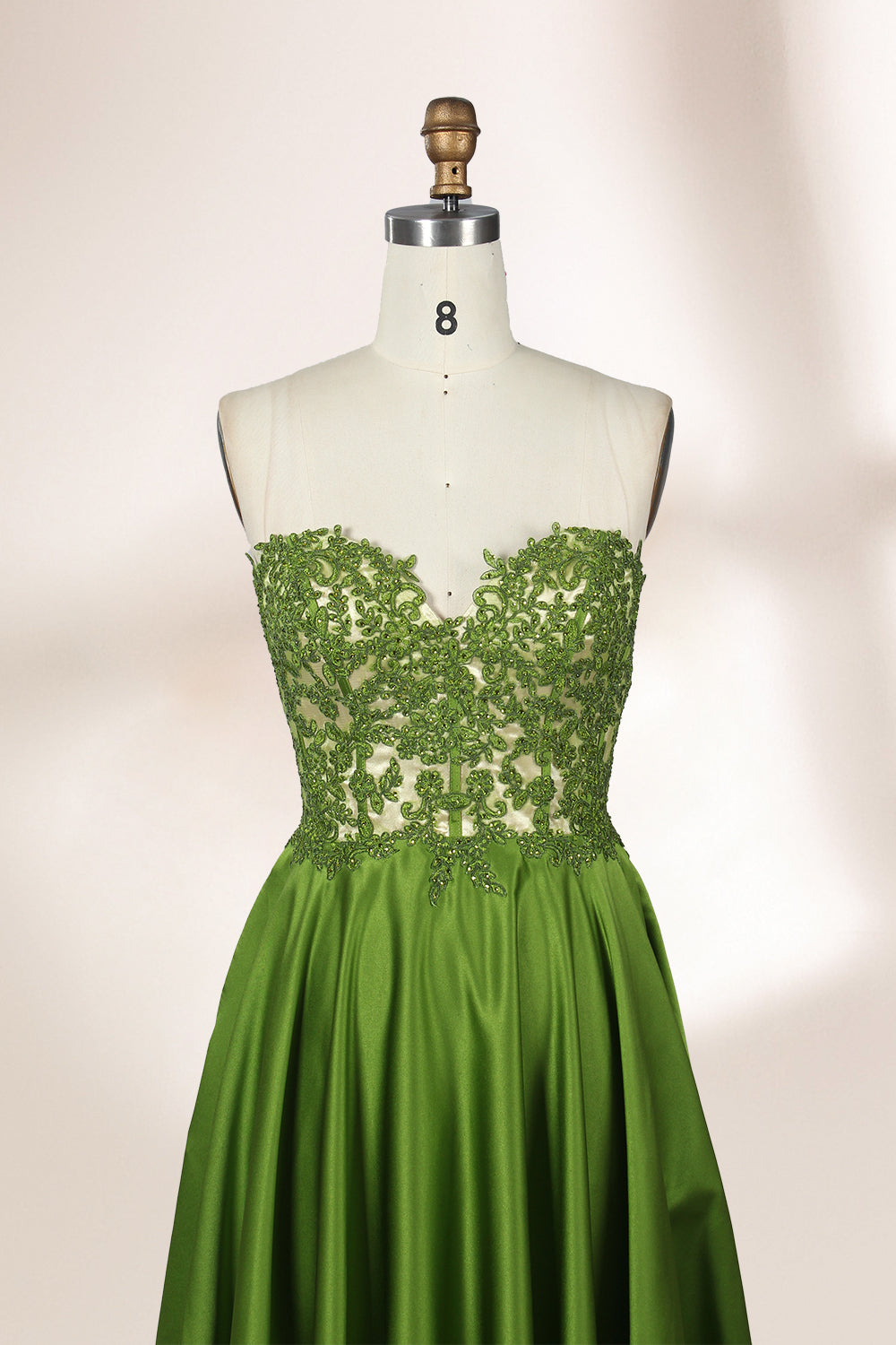 Photograph of an elegant, strapless gown displayed on a mannequin. The gown features a green, satin bodice adorned with intricate lace detailing and a full, floor-length skirt. The background is a soft, neutral gradient, enhancing the gown's vibrant green color. The mannequin is positioned centrally, with the gown's luxurious texture and refined craftsmanship prominently showcased.32639.4