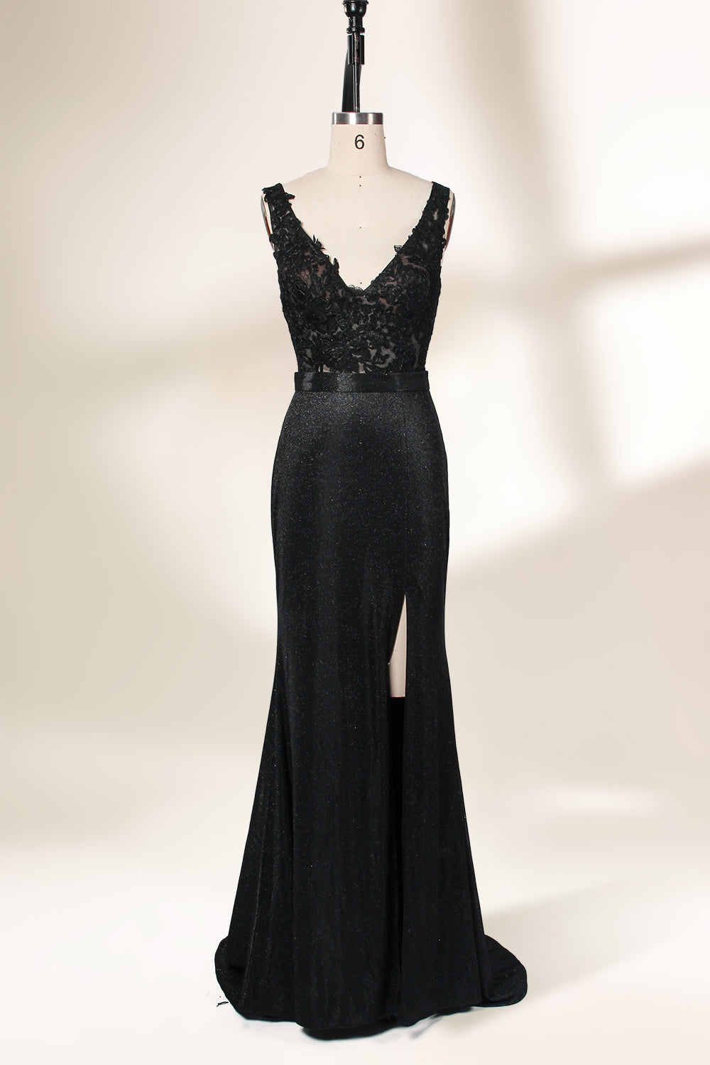 A photograph of a black, floor-length evening gown with a deep V-neck and lace detailing, featuring a thigh-high slit on the left side. The dress is displayed on a white mannequin with a black stand against a soft, beige background with diffused lighting creating gentle shadows. The gown exudes elegance and sophistication.32645.1
