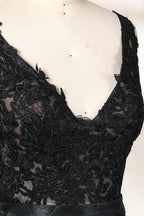 A photograph of a black, floor-length evening gown with a deep V-neck and lace detailing, featuring a thigh-high slit on the left side. The dress is displayed on a white mannequin with a black stand against a soft, beige background with diffused lighting creating gentle shadows. The gown exudes elegance and sophistication.32645.3