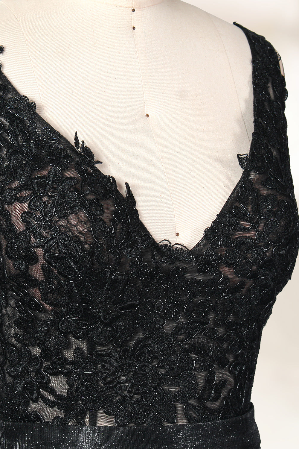 A photograph of a black, floor-length evening gown with a deep V-neck and lace detailing, featuring a thigh-high slit on the left side. The dress is displayed on a white mannequin with a black stand against a soft, beige background with diffused lighting creating gentle shadows. The gown exudes elegance and sophistication.32645.3