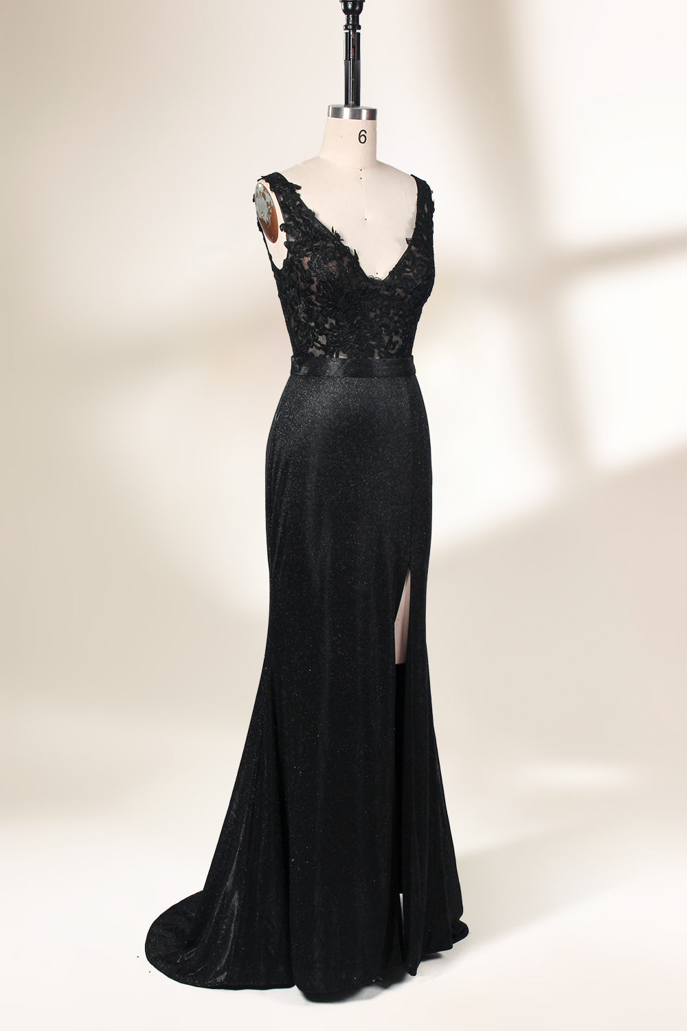 A photograph of a black, floor-length evening gown with a deep V-neck and lace detailing, featuring a thigh-high slit on the left side. The dress is displayed on a white mannequin with a black stand against a soft, beige background with diffused lighting creating gentle shadows. The gown exudes elegance and sophistication.32645.4