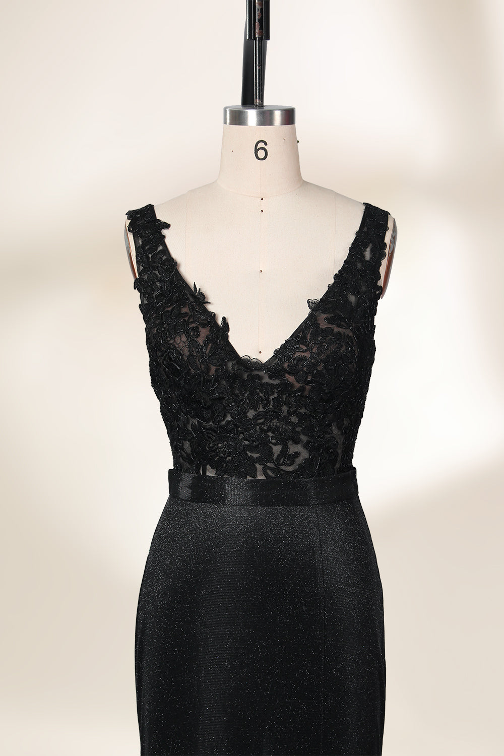 A photograph of a black, floor-length evening gown with a deep V-neck and lace detailing, featuring a thigh-high slit on the left side. The dress is displayed on a white mannequin with a black stand against a soft, beige background with diffused lighting creating gentle shadows. The gown exudes elegance and sophistication.32645.5