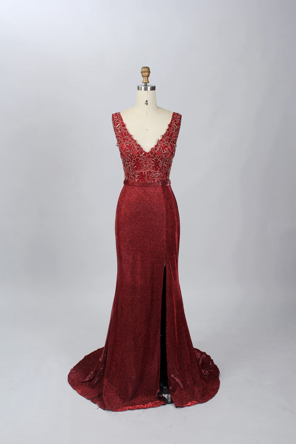 A photograph of a black, floor-length evening gown with a deep V-neck and lace detailing, featuring a thigh-high slit on the left side. The dress is displayed on a white mannequin with a black stand against a soft, beige background with diffused lighting creating gentle shadows. The gown exudes elegance and sophistication.32645.7