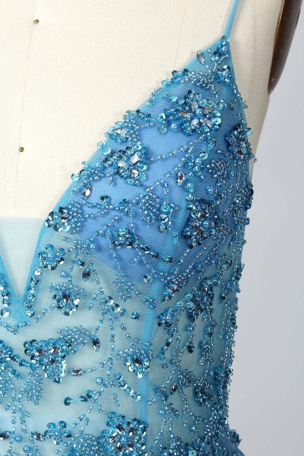 A photograph of a mannequin wearing a floor-length, blue evening gown. The dress features a deep V-neckline adorned with intricate beading, a fitted bodice, and a flowing, high slit skirt. The background is a plain, light beige color, highlighting the gown's elegant design and delicate textures.32656.03