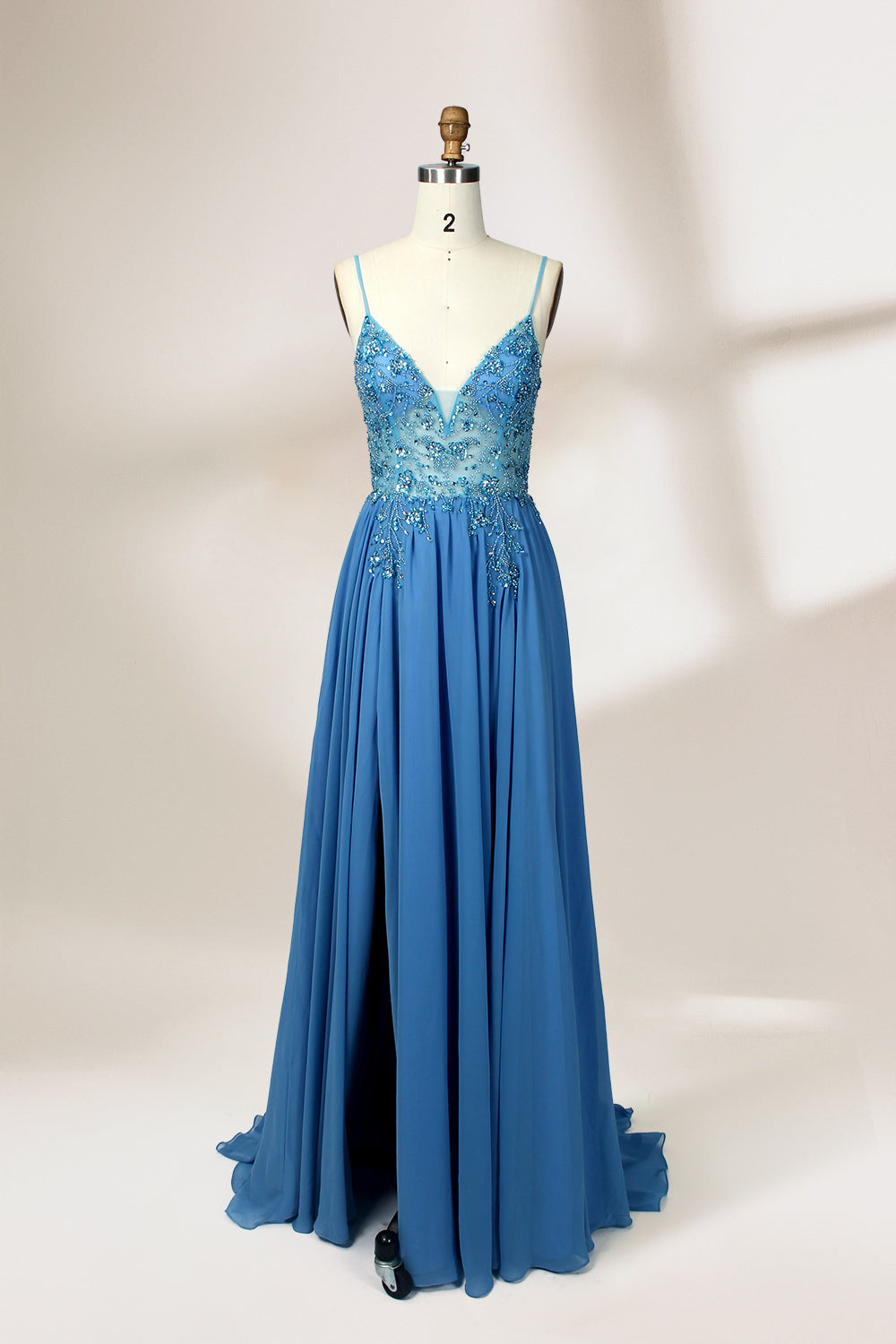 A photograph of a mannequin wearing a floor-length, blue evening gown. The dress features a deep V-neckline adorned with intricate beading, a fitted bodice, and a flowing, high slit skirt. The background is a plain, light beige color, highlighting the gown's elegant design and delicate textures.32656.1