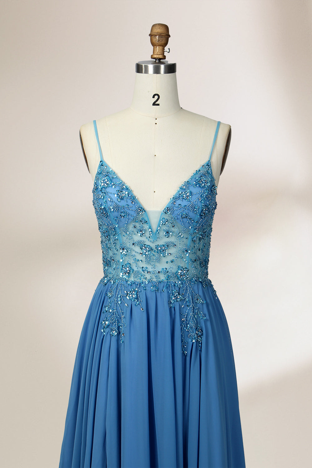 A photograph of a mannequin wearing a floor-length, blue evening gown. The dress features a deep V-neckline adorned with intricate beading, a fitted bodice, and a flowing, high slit skirt. The background is a plain, light beige color, highlighting the gown's elegant design and delicate textures.32656.4
