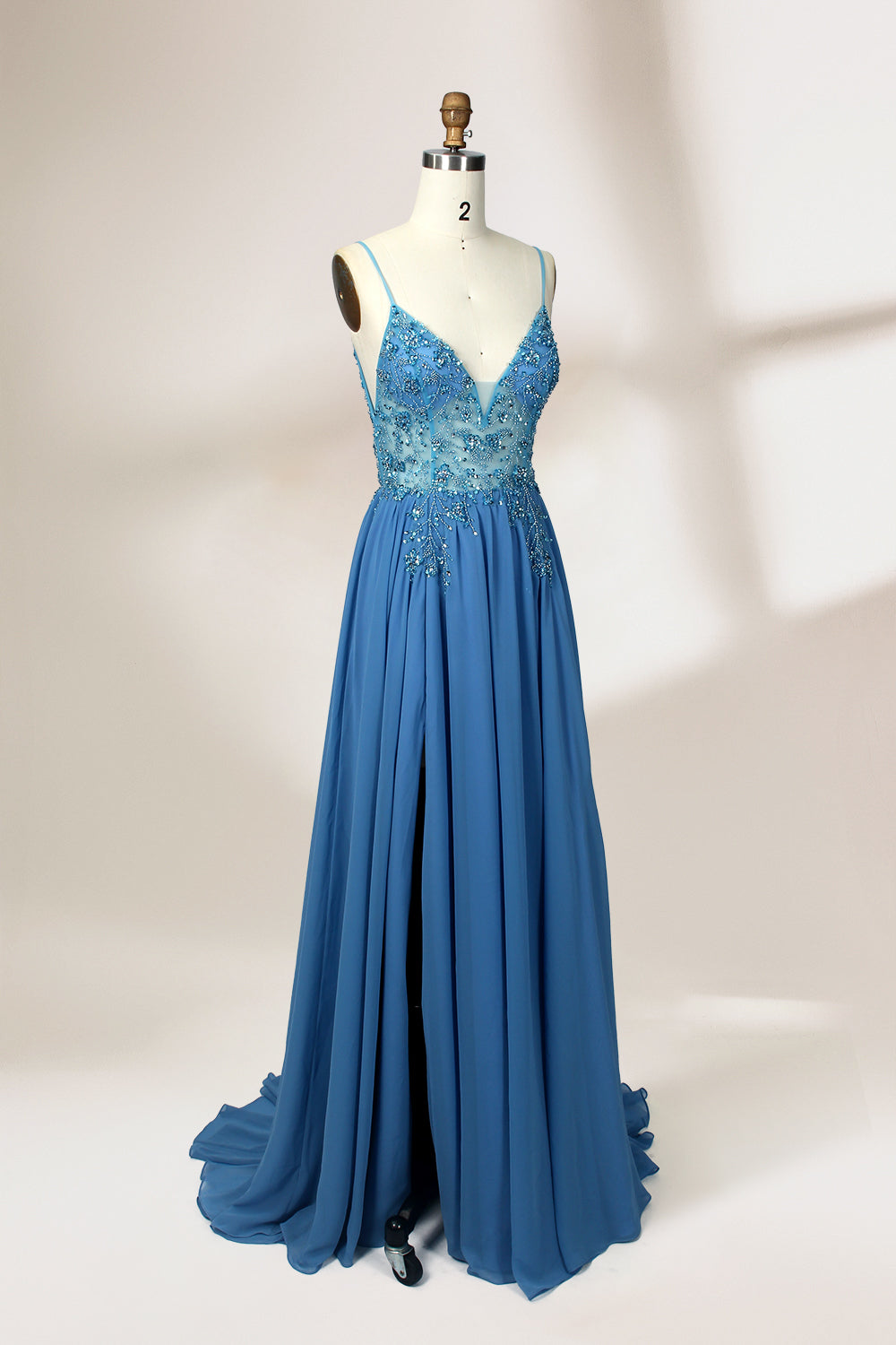 A photograph of a mannequin wearing a floor-length, blue evening gown. The dress features a deep V-neckline adorned with intricate beading, a fitted bodice, and a flowing, high slit skirt. The background is a plain, light beige color, highlighting the gown's elegant design and delicate textures.32656.5