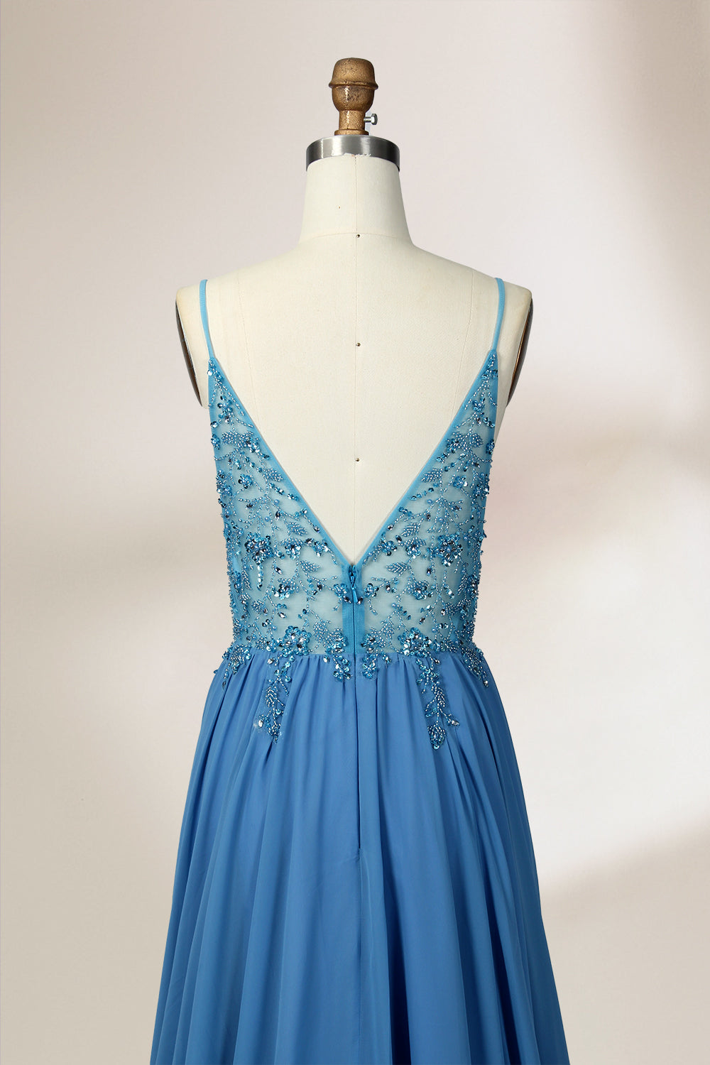 A photograph of a mannequin wearing a floor-length, blue evening gown. The dress features a deep V-neckline adorned with intricate beading, a fitted bodice, and a flowing, high slit skirt. The background is a plain, light beige color, highlighting the gown's elegant design and delicate textures.32656.6