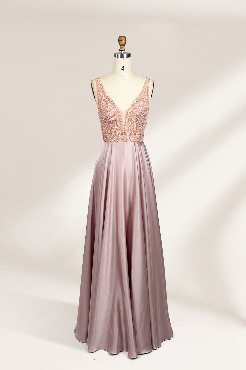 Photograph of a sleeveless, floor-length evening gown displayed on a dress form. The gown features a deep V-neckline adorned with intricate lace detailing and a sheer, blush pink fabric that flows into a smooth, satin skirt. The dress form is positioned against a plain, light beige background, emphasizing the gown's elegant design.01