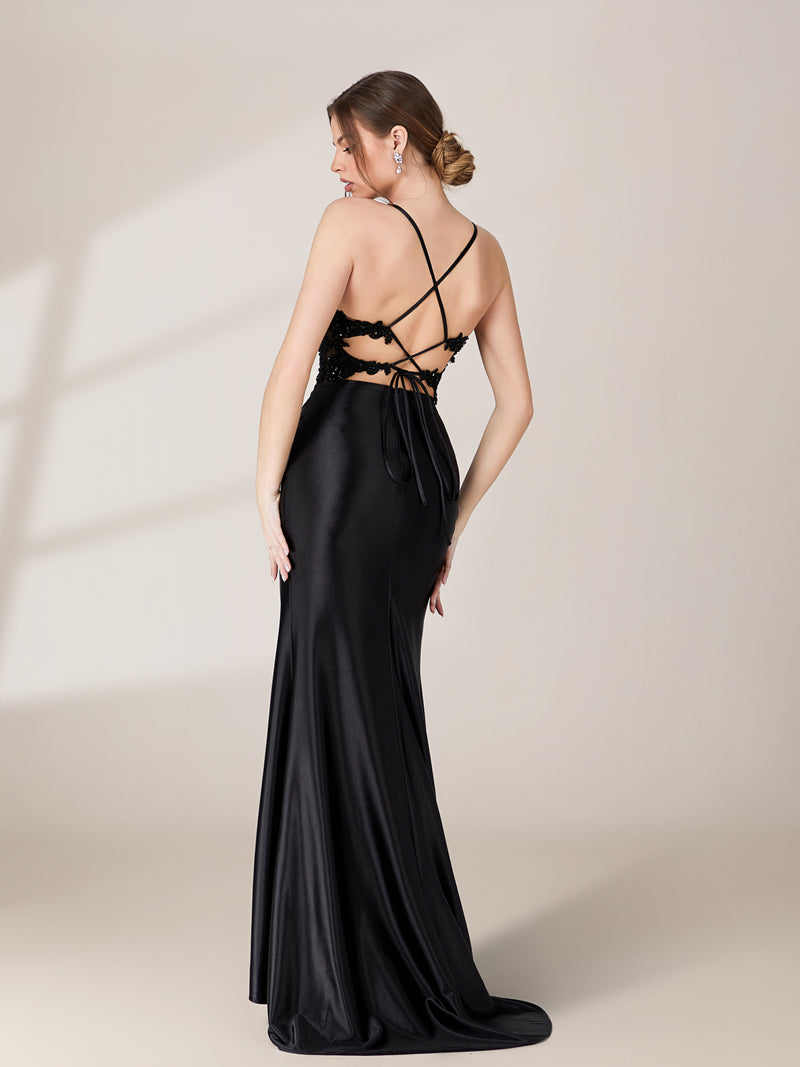 This is a photograph of a young Caucasian woman with light skin, wearing a stunning, floor-length black satin gown with intricate crisscross lacing on the back and a high slit. Her hair is styled in a sleek, low bun. She stands against a plain, light beige background, with soft shadows cast on the wall. The gown's smooth, glossy texture contrasts with her elegant, understated makeup.