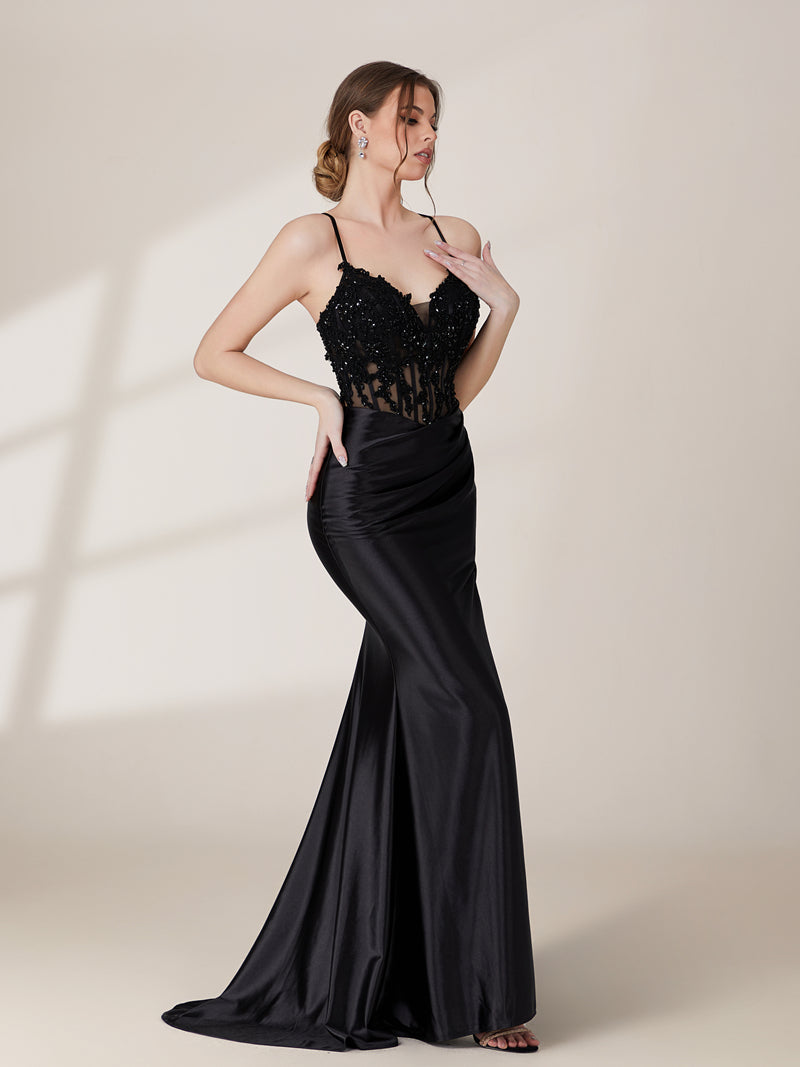 A photograph of a slender, light-skinned woman with long, brown hair in an updo, wearing a black, floor-length evening gown with a deep V-neckline and spaghetti straps. The gown features intricate lace detailing on the bodice and a high slit on the left leg. She accessorizes with a gold ankle bracelet and strappy high heels. The background is a plain, light beige wall with soft shadows cast from a window, creating a minimalist and elegant atmosphere.