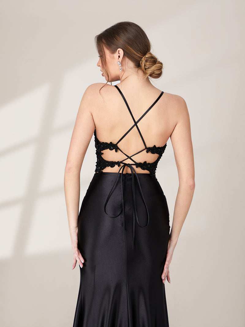 This is a photograph of a young Caucasian woman with light skin, wearing a stunning, floor-length black satin gown with intricate crisscross lacing on the back and a high slit. Her hair is styled in a sleek, low bun. She stands against a plain, light beige background, with soft shadows cast on the wall. The gown's smooth, glossy texture contrasts with her elegant, understated makeup.