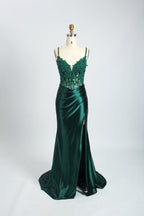 A photograph of a slender, light-skinned woman with long, brown hair in an updo, wearing a green, floor-length evening gown with a deep V-neckline and spaghetti straps. The gown features intricate lace detailing on the bodice and a high slit on the left leg. She accessorizes with a gold ankle bracelet and strappy high heels. The background is a plain, light beige wall with soft shadows cast from a window, creating a minimalist and elegant atmosphere.