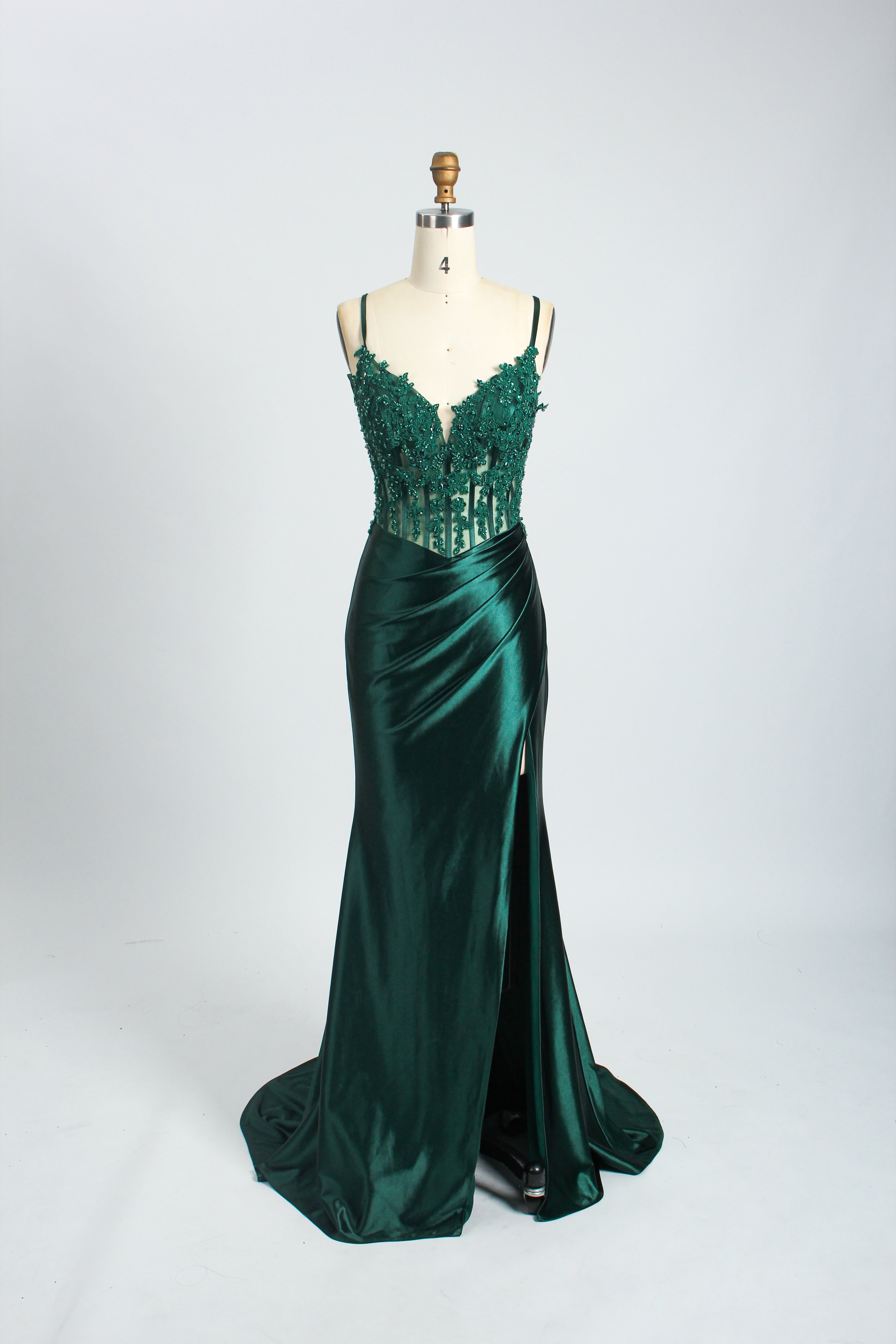 A photograph of a slender, light-skinned woman with long, brown hair in an updo, wearing a green, floor-length evening gown with a deep V-neckline and spaghetti straps. The gown features intricate lace detailing on the bodice and a high slit on the left leg. She accessorizes with a gold ankle bracelet and strappy high heels. The background is a plain, light beige wall with soft shadows cast from a window, creating a minimalist and elegant atmosphere.