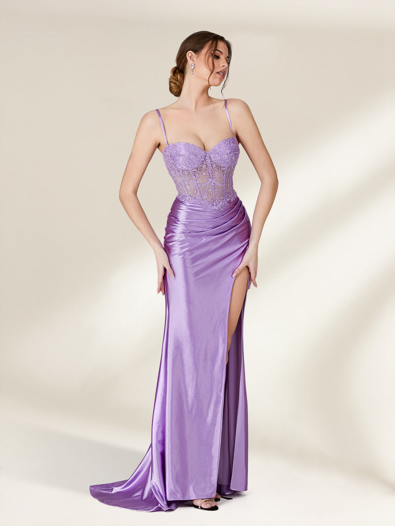 A photograph of a young woman with light skin and brown hair styled in an elegant updo, wearing a shimmering, floor-length, lavender satin gown with a high slit and intricate lace detailing. She stands confidently against a plain, beige background, exuding sophistication and grace.32738-03