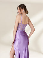 Photograph of a slender, light-skinned woman with brunette hair in a bun, wearing a strapless, lavender satin gown with intricate lace detailing on the back. The gown has a fitted bodice and a flowing, floor-length skirt. She stands against a plain, cream-colored background, with soft shadows adding depth. The gown's texture appears smooth and luxurious. The overall aesthetic is elegant and sophisticated.32738-06
